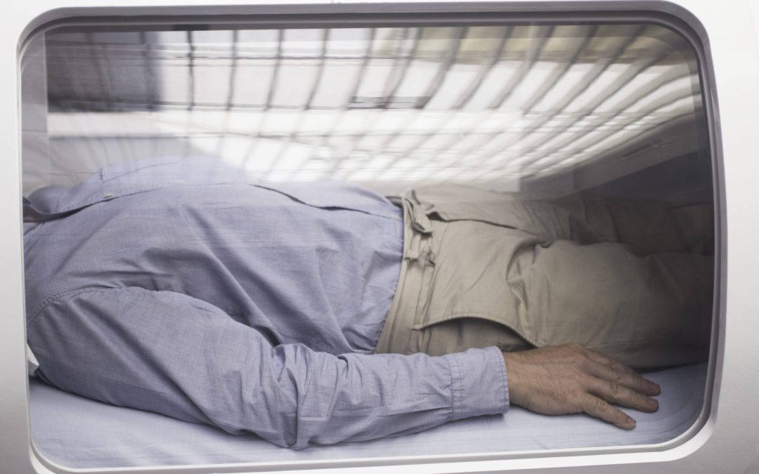 Male patient aged 45-55 wearing blue shirt and beige chino trousers lying down in hyperbaric oxygen chamber receiving Hyperbaric Oxygen Therapy (HBOT) specialised medical treatment for injuries with reflection on glass.
