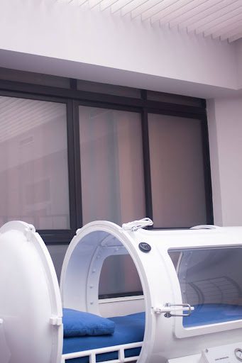 A clean, modern monoplace hyperbaric oxygen therapy chamber with an open hatch, blue pillow, and clear viewing windows inside a professional clinic setting.